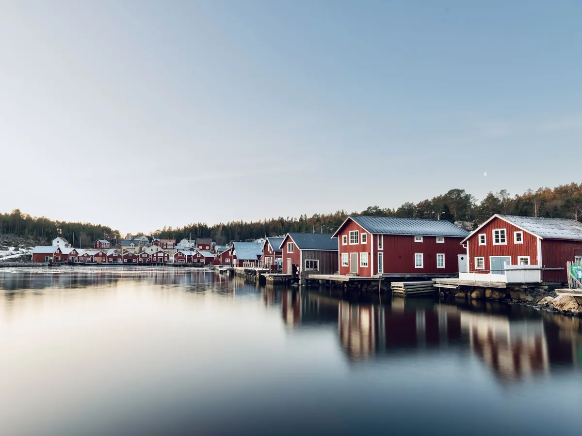 Red wooden boathouses and cottages along calm Nordic water under a pale sky