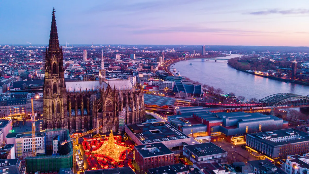 Cologne Cathedral at dusk seen from above, a Christmas market glowing at its foot and the Rhine behind