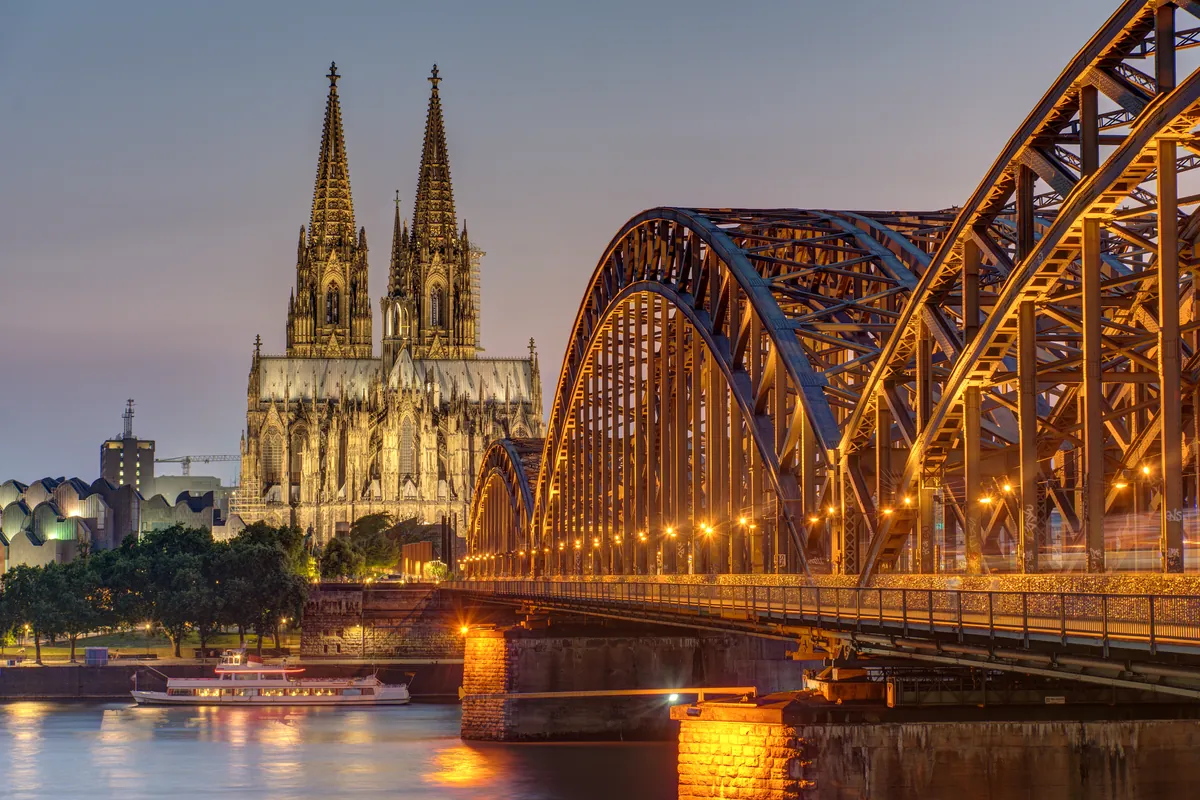 Cologne Cathedral and the lit arches of the Hohenzollern Bridge over the Rhine at dusk, a river cruiser below