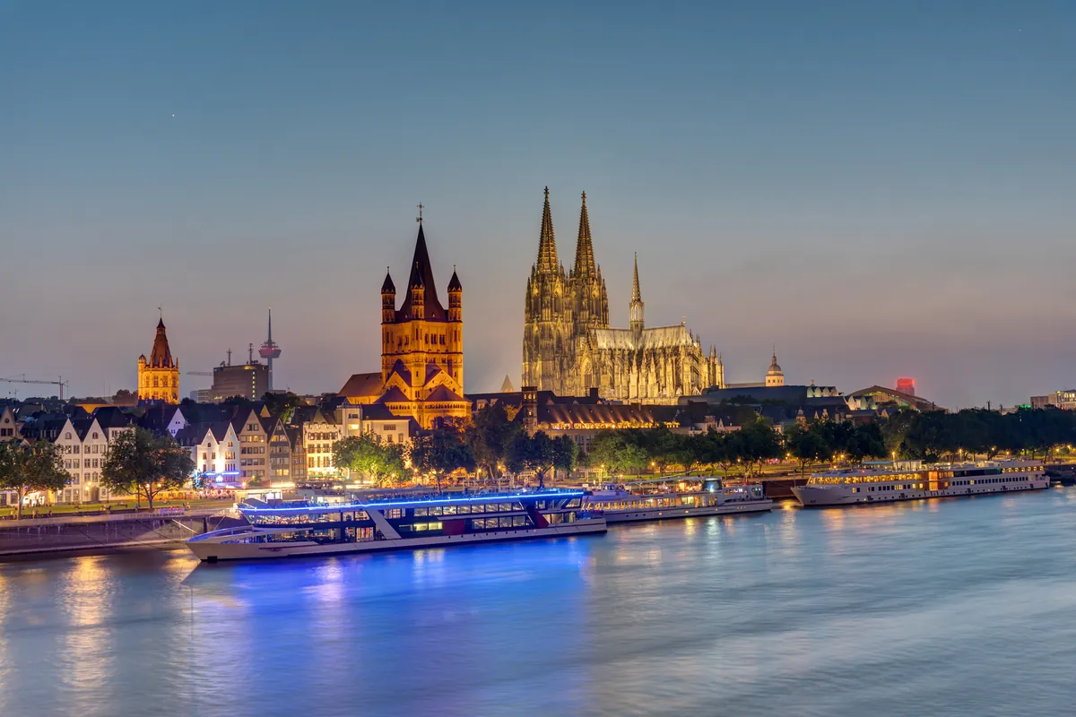 Great St Martin church and Cologne Cathedral above the old town riverside at blue hour, lit boats on the Rhine
