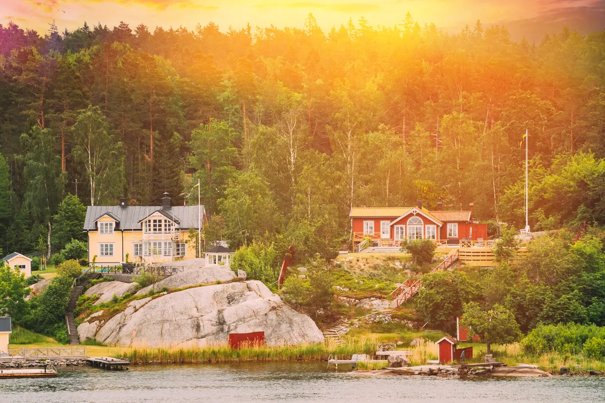 A red wooden house and a yellow villa on a rocky shore by calm water with pine forest behind in warm evening light