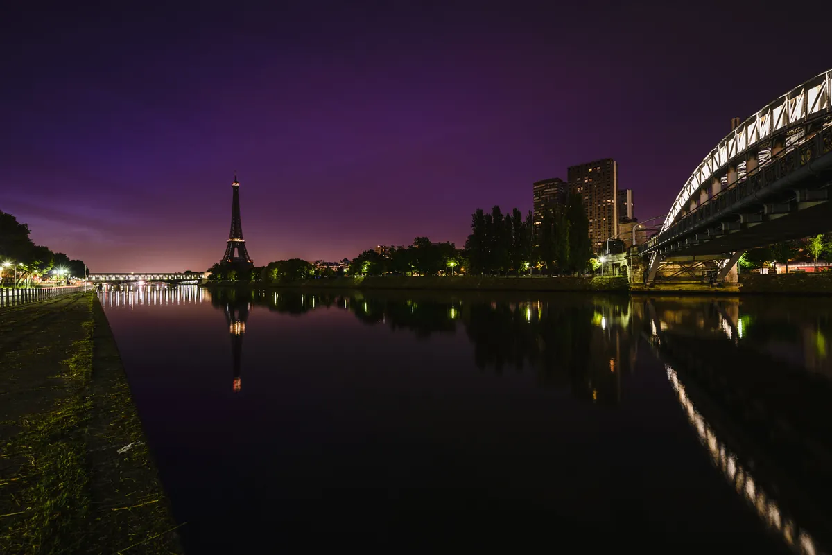 The Seine in Paris at dusk under a violet sky, a lit bridge reflected in the still water and the Eiffel Tower on the horizon