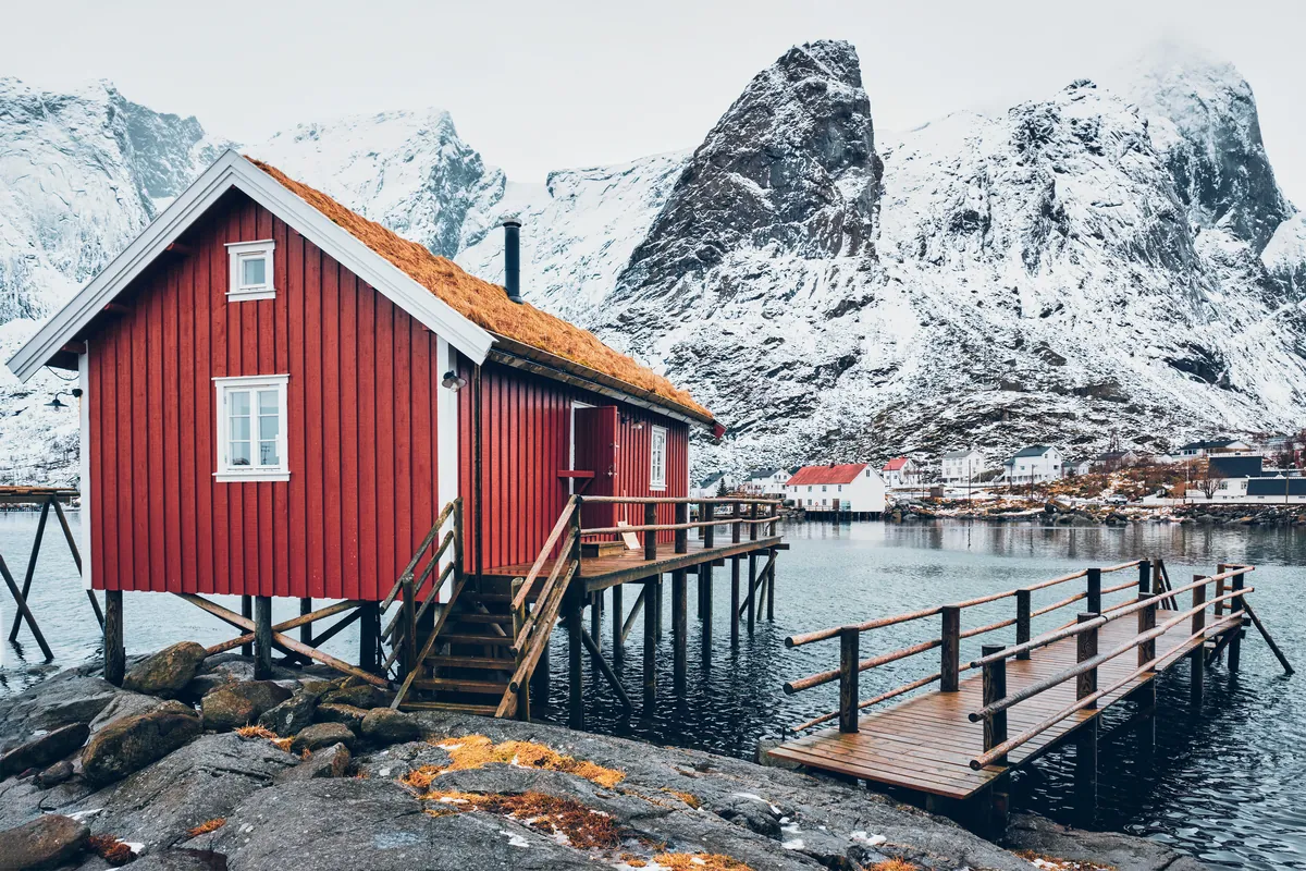 A traditional red rorbu cabin on stilts above the water in Reine on the Lofoten Islands, snowy peaks behind